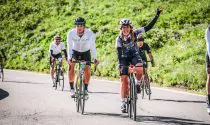 Cyclists riding near green fields on open roads, raising their hands and smiling for a photo.