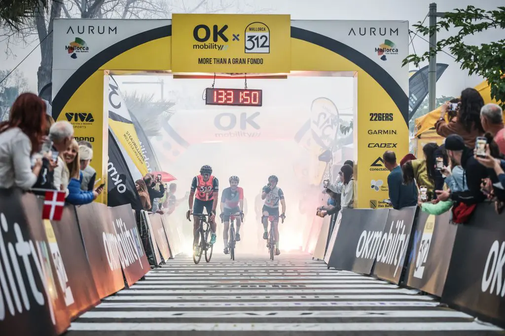 Mallorca 312 event finish line, showing spectators cheering as cyclists appear through a cloud of smoke towards the finish line. 