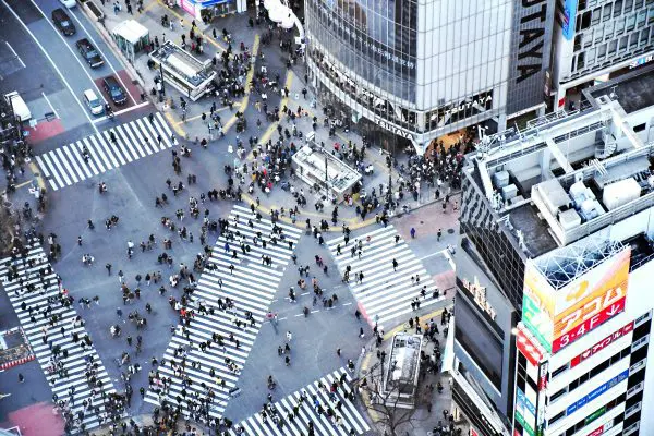 A high-angle view of Shibuya Crossing in Tokyo, filled with crowds of people walking in all directions across the intersection, surrounded by tall buildings with bright neon signs and digital billboards.