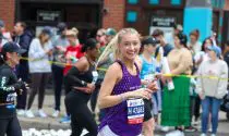 A women, taking part in a marathon running in a purple running vest. Views of spectators and other runners in the background.