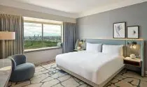 A hotel room, with a double bed, white bedding, picture wall art on grey walls and a window with views of green trees and fields.