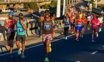 An action shot of a Sanlam Cape Town Marathon runner racing along a city highway, smiling with both arms out, creating the peace sign gesture on both hands.