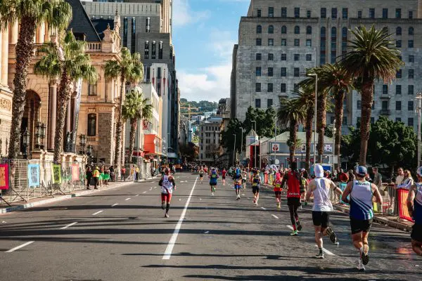 Runners participating in the Sanlam Cape Town Marathon running through the city with palm trees on the right hand side of the road, and buildings in the background