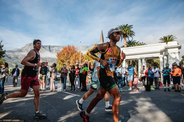 Two runners participating in the Sanlam Cape Town Marathon running with the crowd in the background with Mount Nelson Hotel white marble archway behind