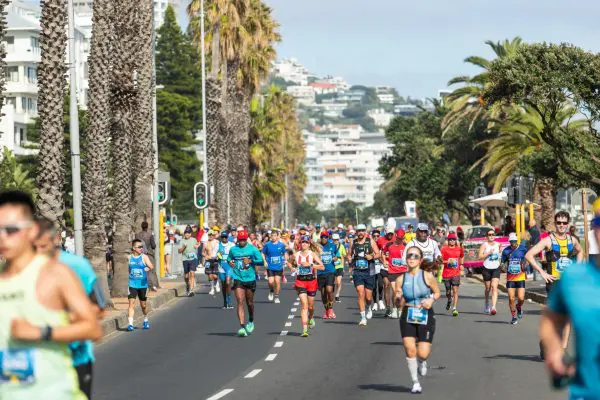 Runners participating in the Sanlam Cape Town Marathon running down a road with houses on the hills in the background and large trees either side of the road