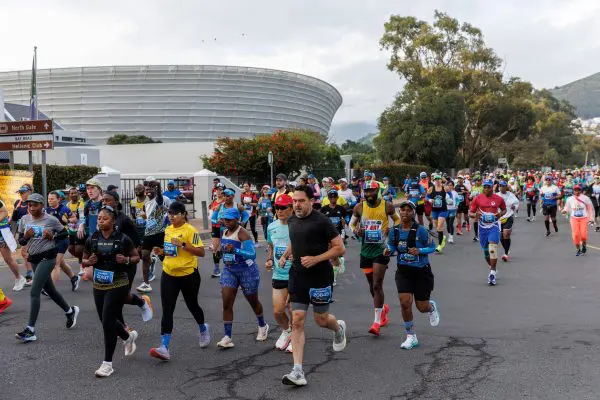 Runners participating in the Sanlam Cape Town Marathon running on the road with a stadium in the background, grey sky and green trees behind