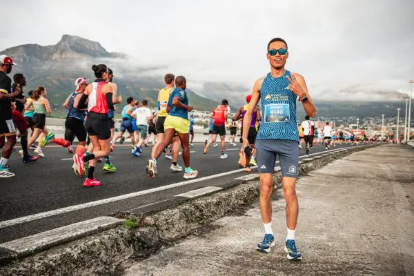 Runners participating in the Sanlam Cape Town Marathon in the background, in the foreground a runner standing still with blue sunglasses on, holding up two fingers and smiling