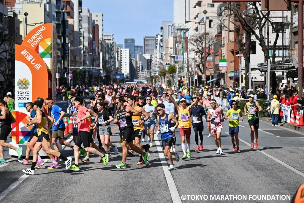 Runners approaching a corner turning point in the Tokyo Marathon, with one runner facing the camera with his arms raised in the air.