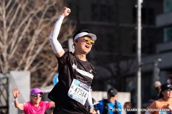 Runner at the Toyko Marathon crossing the finish line with his arm raised in the air with a clenched fist and smiling, celebrating his race.
