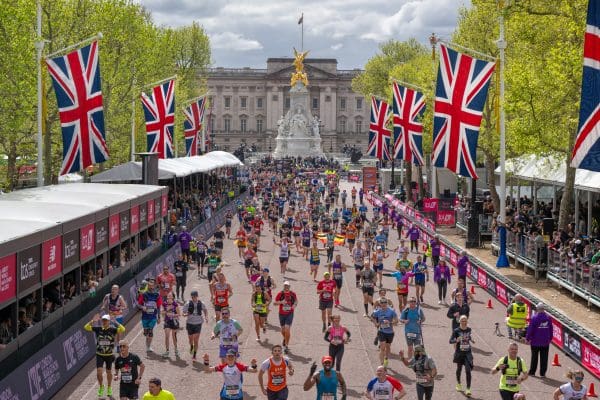 Runners participating in London Marathon near Buckingham Palace in London, with Union Jack flags lining the street and the Victoria Memorial in the background. Spectators are watching from stands on both sides of the finish line