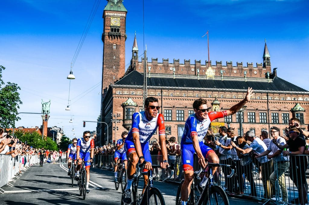 Tour de France cyclists waving to crowds of spectators under a blue sky and smooth roads. 